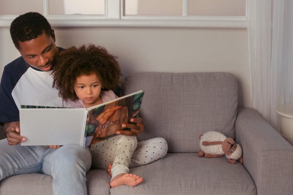 Pai sentado com a filha, lendo no sofá. Imagem ilustrativa texto literatura aproxima família.