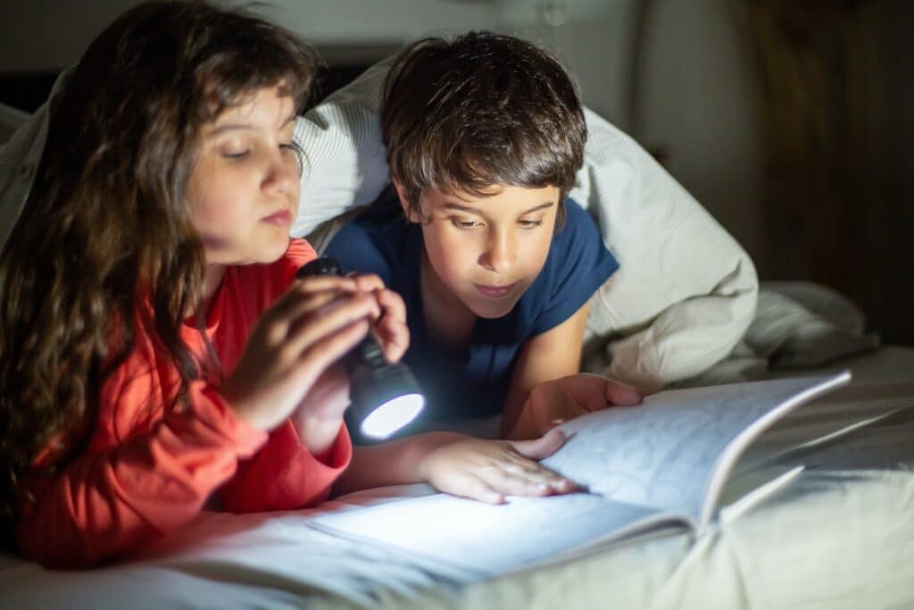 Menina e menino debaixo de cobertas, com lanterna, lendo no escuro. Imagem ilustrativa texto leitura desenvolve.